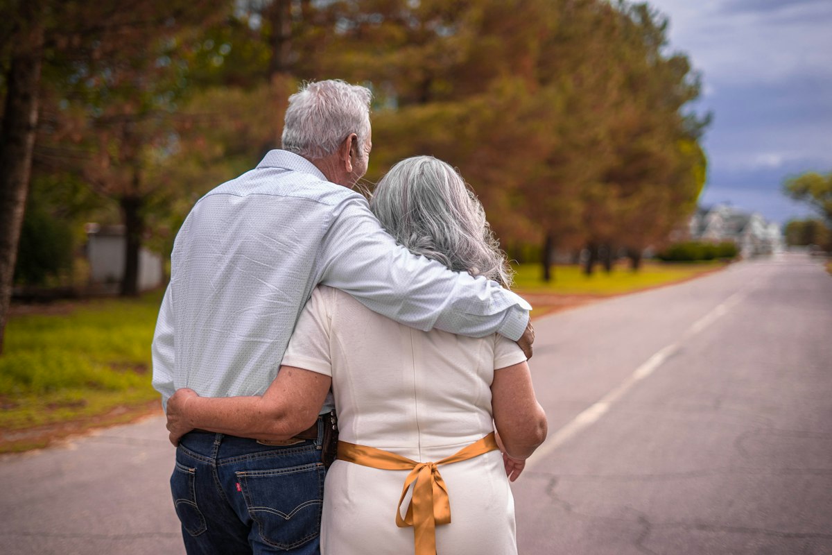 Un couple aux cheveux gris marchant bras dessus bras dessous sur une route bordée d'arbres